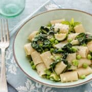 Un primo piano di pasta di verdure verdi in un piatto verde chiaro su lino azzurro. Il piatto mostra spinaci, piselli e zucchine