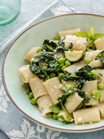 Un primo piano di pasta di verdure verdi in un piatto verde chiaro su lino azzurro. Il piatto mostra spinaci, piselli e zucchine
