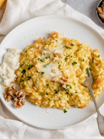Una vista dall'alto del risotto alla zucca con gorgonzola in un piatto bianco su un lino luminoso. È condito con prezzemolo fresco e noci
